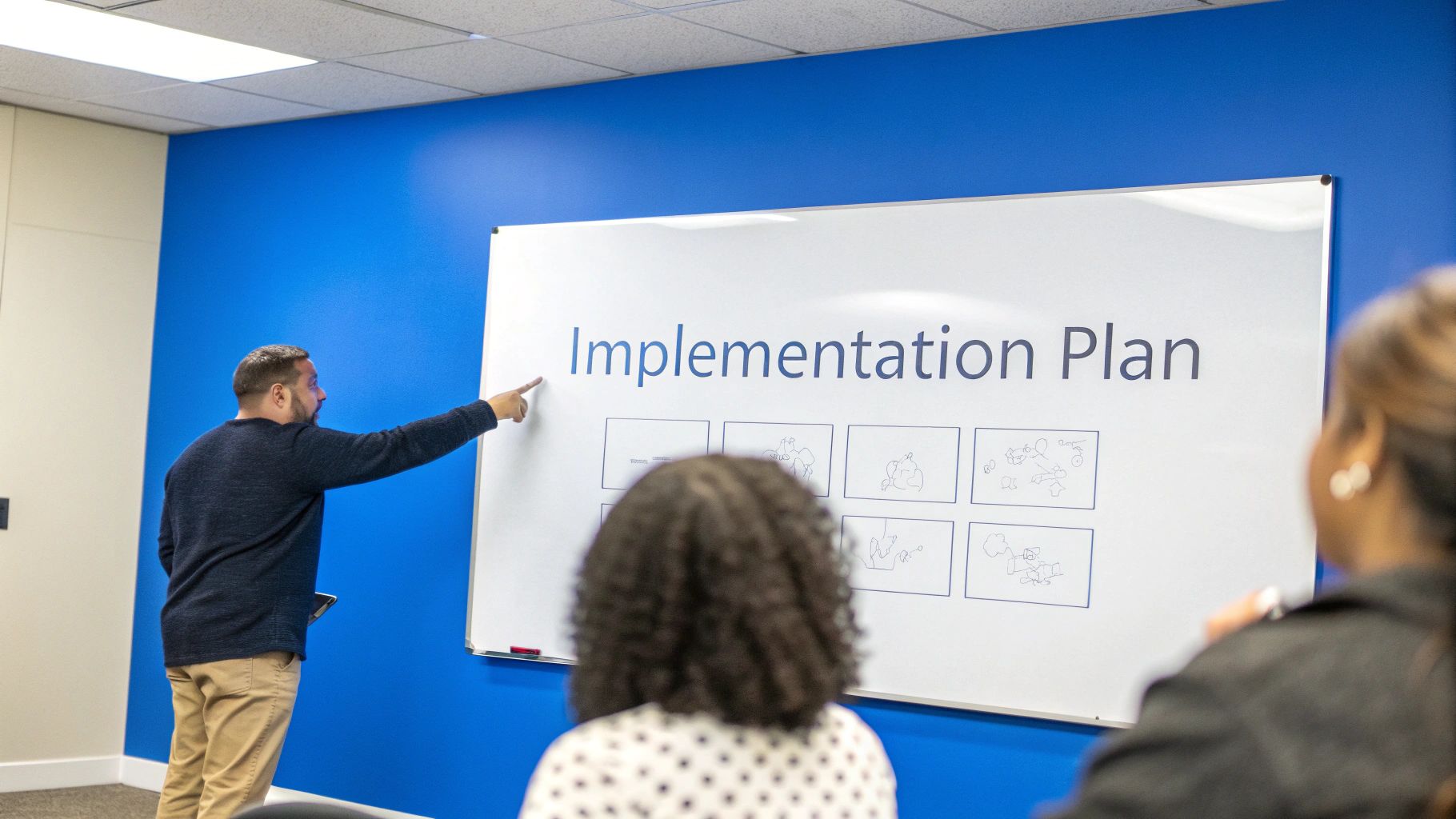 A man points at a whiteboard displaying 'Implementation Plan' during a presentation to a team.